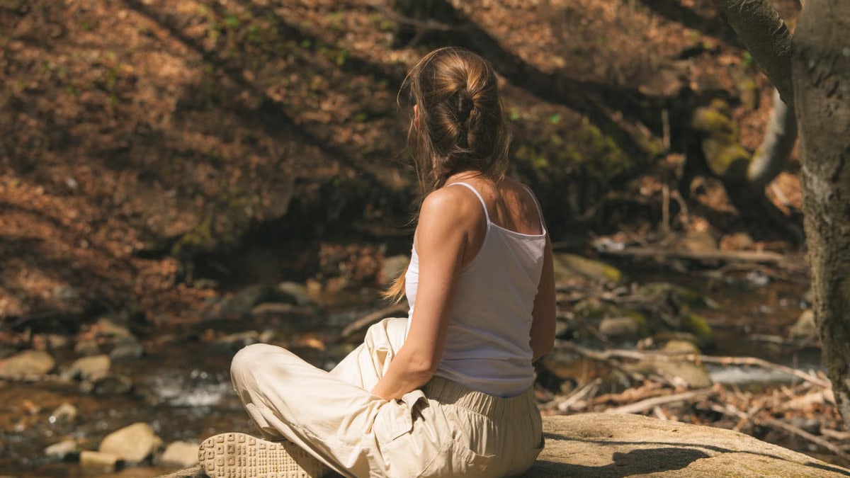 A woman in a white tank top sits peacefully in a forest, enjoying nature.
